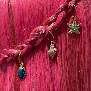 May include: Three gold hair rings with charms. The charms are a green starfish, a silver seashell, and a blue seashell. The hair rings are in a braid of pink hair.