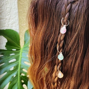 May include: A close-up of a braid with three seashell hair charms. The charms are attached to small silver hoops. The seashells are pink, light green, and gold with a pearl in the center. The hair is brown with reddish highlights.