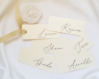 May include: Cream-colored place cards with elegant calligraphy names, including Lauren, Karina, and others. A cream-colored ribbon and a fabric flower accent the cards, arranged on a white fabric surface. Ideal for weddings or special events.