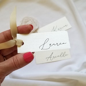 May include: Three white place cards with calligraphy names written in black ink. The names are "Lauren", "Arielle", and "Keren". A hand is holding a place card with the name "Lauren" and a beige ribbon.