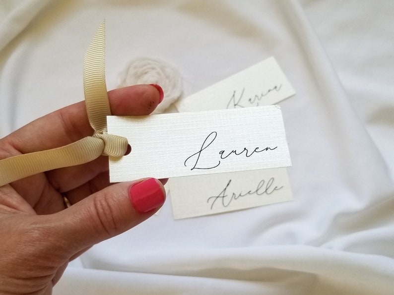 May include: Three white place cards with calligraphy names written in black ink. The names are "Lauren", "Arielle", and "Keren". A hand is holding a place card with the name "Lauren" and a beige ribbon.