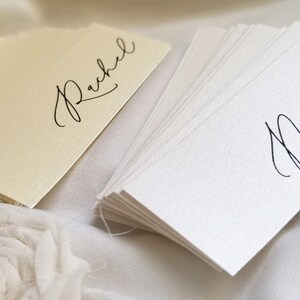 May include: A stack of blank white place cards with the name "Natal" written in black calligraphy. A single place card with the name "Rachel" written in black calligraphy is on a cream background.