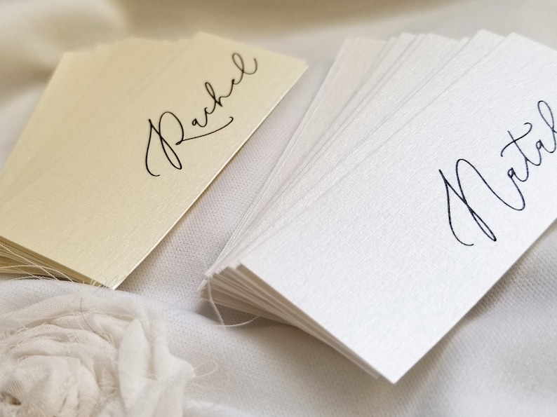 May include: A stack of blank white place cards with the name "Natal" written in black calligraphy. A single place card with the name "Rachel" written in black calligraphy is on a cream background.