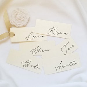 May include: Six white place cards with black calligraphy names: Lauren, Kerina, Shannon, Terra, Girda, and Arielle. The cards are arranged on a white fabric background.