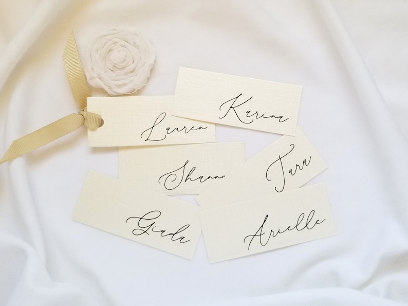 May include: Six white place cards with black calligraphy names: Lauren, Kerina, Shannon, Terra, Girda, and Arielle. The cards are arranged on a white fabric background.