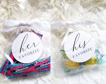 Op de afbeelding: Twee doorzichtige plastic zakjes die met een wit lint zijn vastgebonden, elk met een selectie snoepjes. Eén zakje heeft een witte ronde sticker met de tekst "haar favoriet" in zwarte letters. Het andere zakje heeft een witte ronde sticker met de tekst "zijn favoriet" in zwarte letters.