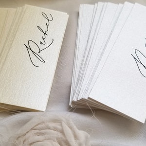May include: Two stacks of place cards, one ivory and one white, with the names "Rachel" and "Natalie" written in black calligraphy.