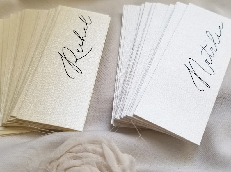 May include: Two stacks of place cards, one ivory and one white, with the names "Rachel" and "Natalie" written in black calligraphy.