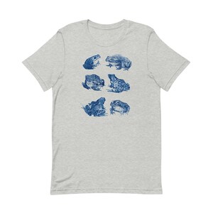 K&ouml;nnte beinhalten: Ein hellgraues T-Shirt mit einem blauen Aufdruck von verschiedenen Fr&ouml;schen in verschiedenen Posen.