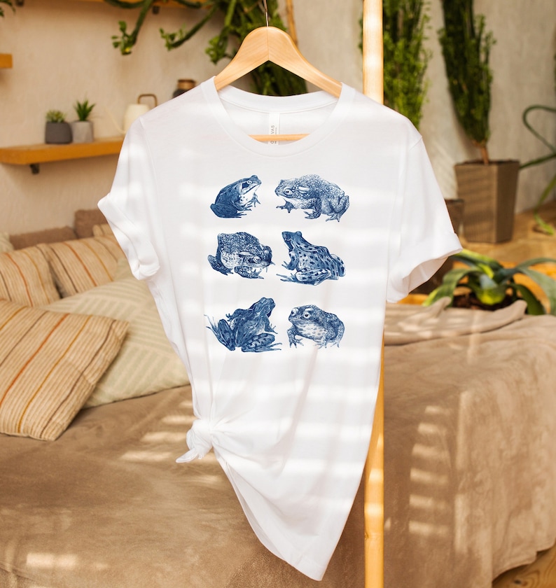 K&ouml;nnte beinhalten: Wei&szlig;es T-Shirt mit einem blauen Aufdruck von sechs Fr&ouml;schen in verschiedenen Posen.