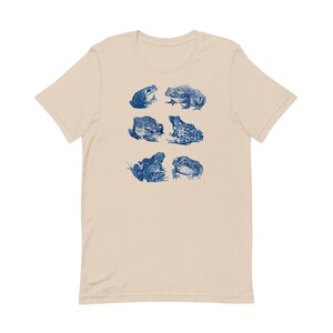 K&ouml;nnte beinhalten: Ein beiges T-Shirt mit einem blauen Aufdruck von verschiedenen Fr&ouml;schen in verschiedenen Posen.