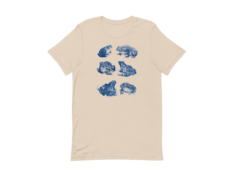K&ouml;nnte beinhalten: Ein beiges T-Shirt mit einem blauen Aufdruck von verschiedenen Fr&ouml;schen in verschiedenen Posen.
