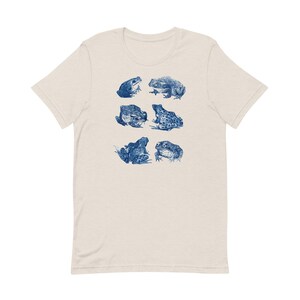 K&ouml;nnte beinhalten: Ein hellbeiges T-Shirt mit einem blauen Aufdruck von sechs Fr&ouml;schen in verschiedenen Posen.