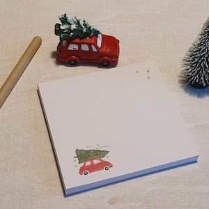 Peut inclure: Bloc-notes blanc avec une illustration de voiture rouge transportant un sapin de Noël et le texte "Merry Christmas". Une figurine de petite voiture rouge avec un arbre sur le dessus, un petit sapin de Noël et un stylo en bois sont également présents.