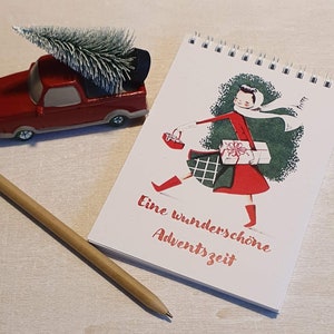 Könnte beinhalten: Ein roter Spielzeug-Pick-up-Truck mit einem kleinen Weihnachtsbaum neben einem Spiralnotizblock mit einer festlichen Illustration. Das Notizbuch zeigt eine Frau in rotem Mantel und Schal, die Geschenke trägt, mit dem Text "Eine wunderschöne Adventszeit". Ein Bleistift aus Holz ist ebenfalls vorhanden.