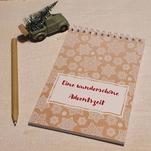 May include: A spiral-bound notebook with a snowflake pattern and the text "Eine wunderschöne Adventszeit" in red. A small green truck carrying a Christmas tree and a pen are also in the image.