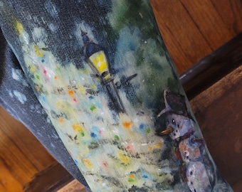 May include: Close-up of denim pants featuring a painted winter scene. The artwork depicts a lamppost, a snowman, and colorful lights against a dark background. The painting style is impressionistic, with visible brushstrokes and a soft color palette.