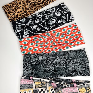 May include: Set of five Halloween-themed headbands. The headbands feature different patterns, including leopard print, skulls and bones, eyeballs, spiderwebs, and a patchwork design with ghosts, pumpkins, and checkerboard patterns. The headbands are made of a stretchy fabric and are designed to be worn as a headband or hair tie.