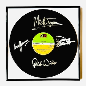 May include: A framed black vinyl record featuring the Atlantic label for the song I Want to Know What Love Is by Foreigner. The record surface displays four distinct silver ink signatures from the band members positioned around the center label.
