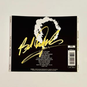 May include: A Bob Dylan Greatest Hits CD case with a black background. The album title and artist name are in white, with gold lettering for the signature. Song titles are listed on the back.