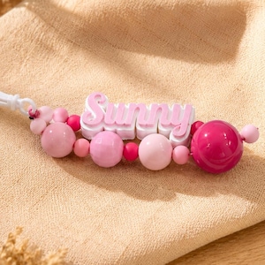 Peut inclure: Un porte-clés composé d'un clip en plastique blanc attaché à une série de perles rondes dans des tons variés de rose et de magenta. Un bloc rectangulaire blanc central affiche le nom Sunny en lettres roses en relief et en 3D.