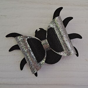 May include: A black and silver glitter spider bow. The bow is shaped like a spider with eight legs and a bow in the center.