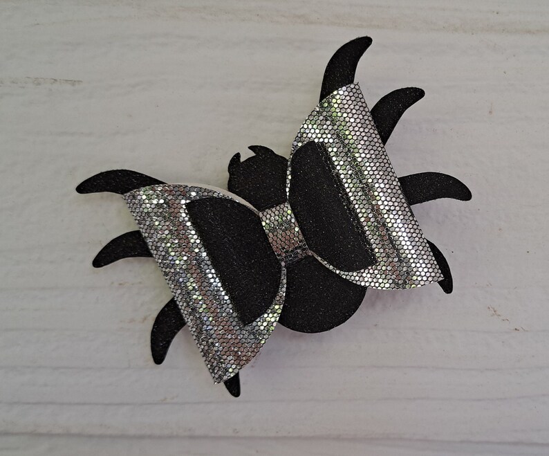 May include: A black and silver glitter spider bow. The bow is shaped like a spider with eight legs and a bow in the center.