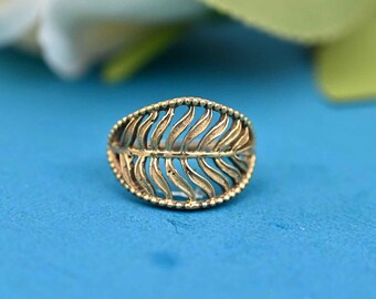 May include: A gold-colored ring with a leaf design. The ring features a detailed, openwork pattern of curved lines resembling leaf veins, surrounded by a beaded border. The ring is set against a blue background.