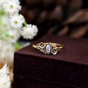 May include: A gold ring featuring a white gemstone and floral details. The ring is displayed on a brown velvet surface.