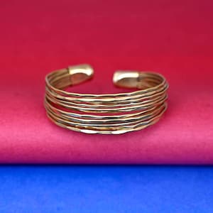 May include: A gold-toned bangle bracelet made of multiple thin bands, each with a hammered texture. The bracelet is open and has a slightly curved shape.
