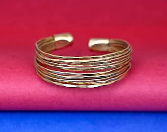 May include: A gold-toned bangle bracelet made of multiple thin bands, each with a hammered texture. The bracelet is open and has a slightly curved shape.