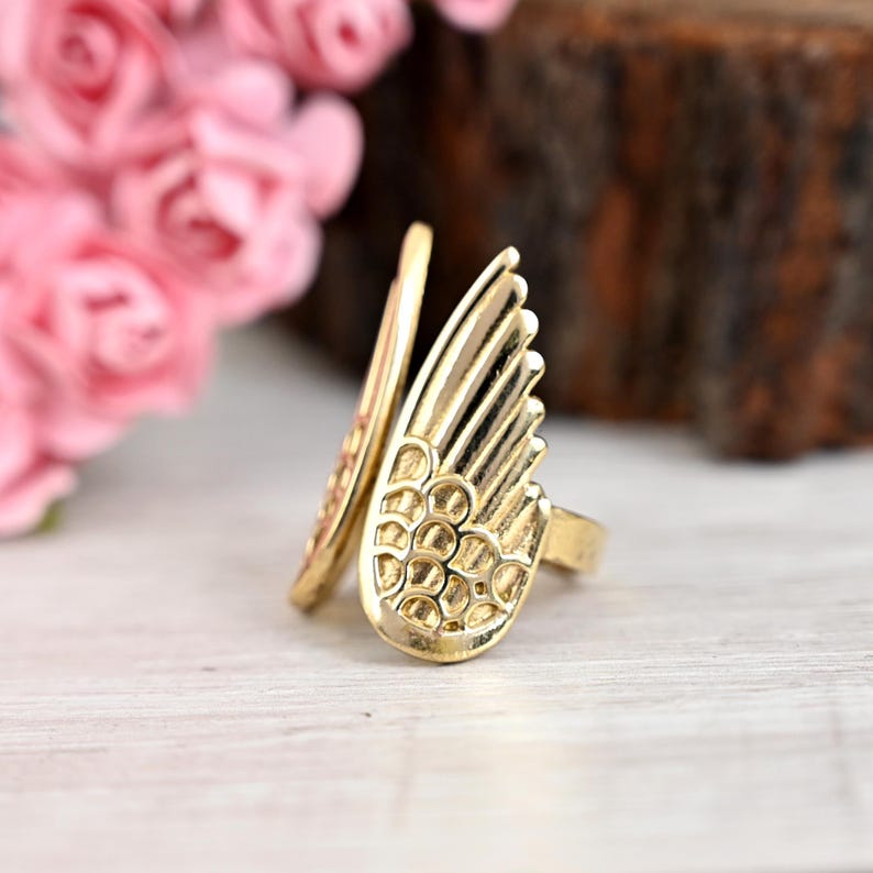 K&ouml;nnte beinhalten: Ein goldfarbener Ring mit einem Fl&uuml;geldesign. Der Ring hat eine detaillierte Fl&uuml;gelform mit strukturierten Mustern und einem glatten, gebogenen Band. Der Ring ist vor einem verschwommenen Hintergrund mit rosa Blumen und einem Holzelement platziert.