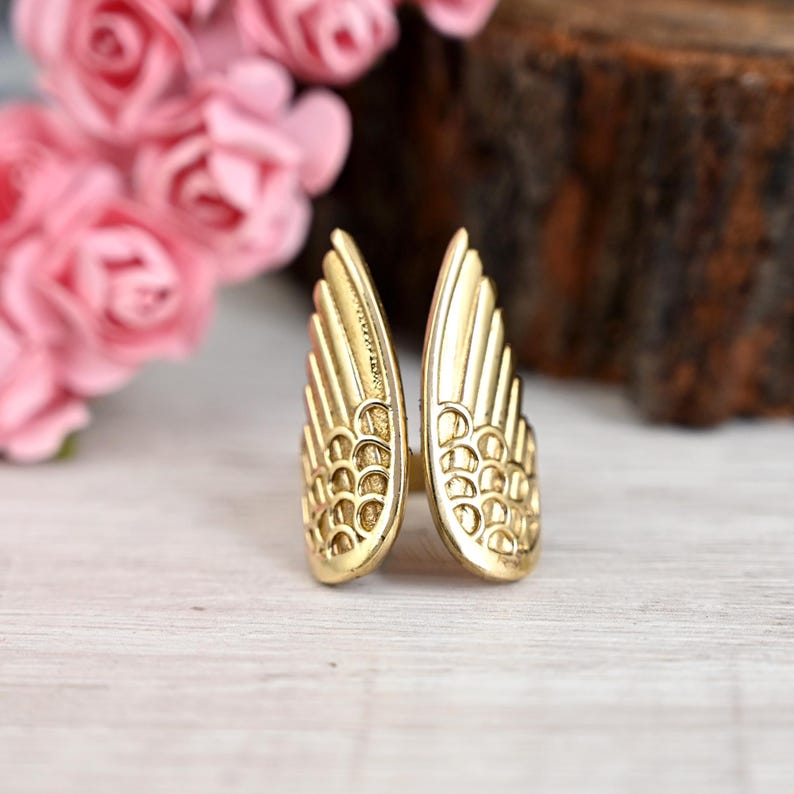 K&ouml;nnte beinhalten: Ein goldfarbener Ring in Form von Engelsfl&uuml;geln. Der Ring weist detaillierte Federstrukturen auf und ist vor einem verschwommenen Hintergrund aus rosa Blumen und einer Holzoberfl&auml;che platziert. Der Ring ist ein Schmuckst&uuml;ck.