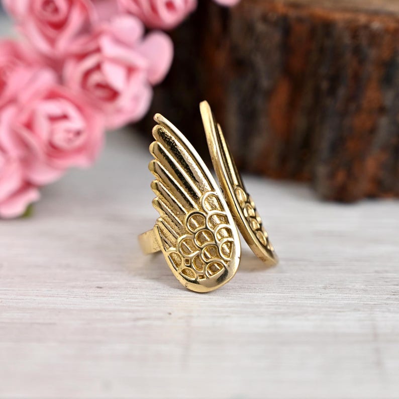 K&ouml;nnte beinhalten: Ein goldfarbener Ring mit einem Fl&uuml;geldesign. Der Ring weist detaillierte, federartige Muster auf und ist vor einem verschwommenen Hintergrund mit rosa Blumen und einem Holzelement platziert. Das Design des Rings erinnert an Engelsfl&uuml;gel.