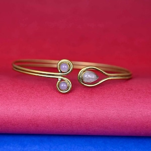 May include: A gold-toned cuff bracelet with two light purple circular accents and a teardrop-shaped, light-colored stone. The bracelet is set against a red and pink background.