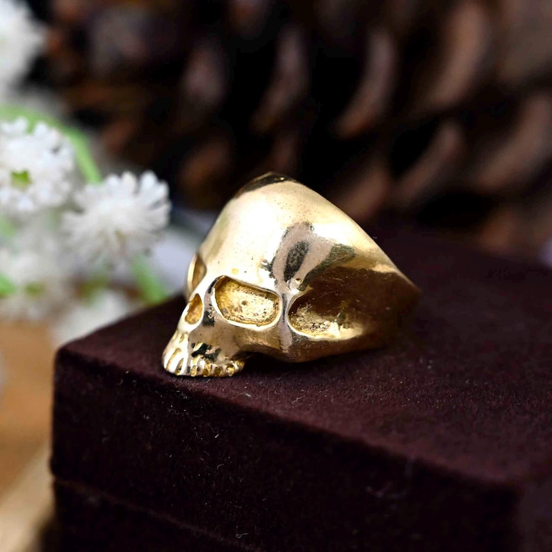 May include: A gold skull ring with a detailed design. The ring is sitting on a brown velvet box.
