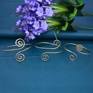 May include: Three gold-colored arm cuffs with spiral and swirl designs. The cuffs are displayed on a blue surface, with purple flowers in the background. The arm cuffs have a delicate, elegant appearance.