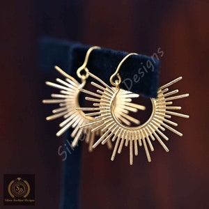 May include: Gold-toned earrings with a sunburst design. The earrings are made of metal and have a hoop shape with a circle of spikes radiating outwards.