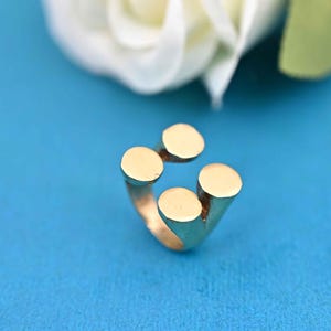 May include: A gold-colored ring with four raised, circular accents. The ring is set against a bright blue background, with a blurred white flower in the upper portion of the image. The ring's design is modern and minimalist.