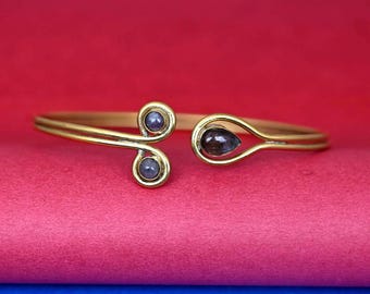 May include: A gold-toned bangle bracelet with three dark purple gemstones. The bracelet is open and has a curved design.
