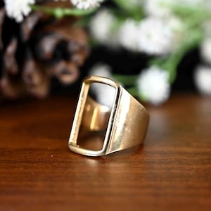 May include: A gold-colored ring with a rectangular opening in the center. The ring is set on a dark wooden surface, with blurred white flowers in the background. The ring's design is simple and modern.