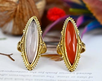 May include: Two gold rings with oval-shaped gemstones. The ring on the left has a white gemstone, and the ring on the right has a red gemstone.