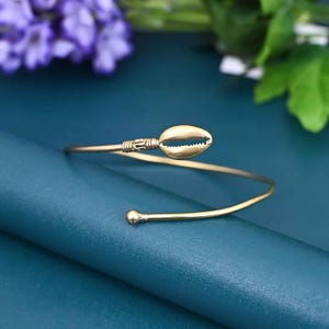 May include: A gold-colored arm cuff bracelet featuring a seashell design. The cuff has a spiral shape with a small ball on one end and a seashell on the other. The bracelet is set against a teal background with purple flowers.