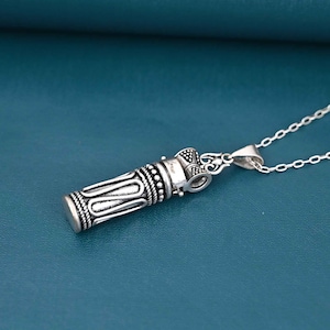 May include: A silver pendant necklace with a cylindrical design. The pendant has detailed patterns and hangs from a fine chain. The background is a teal colour.