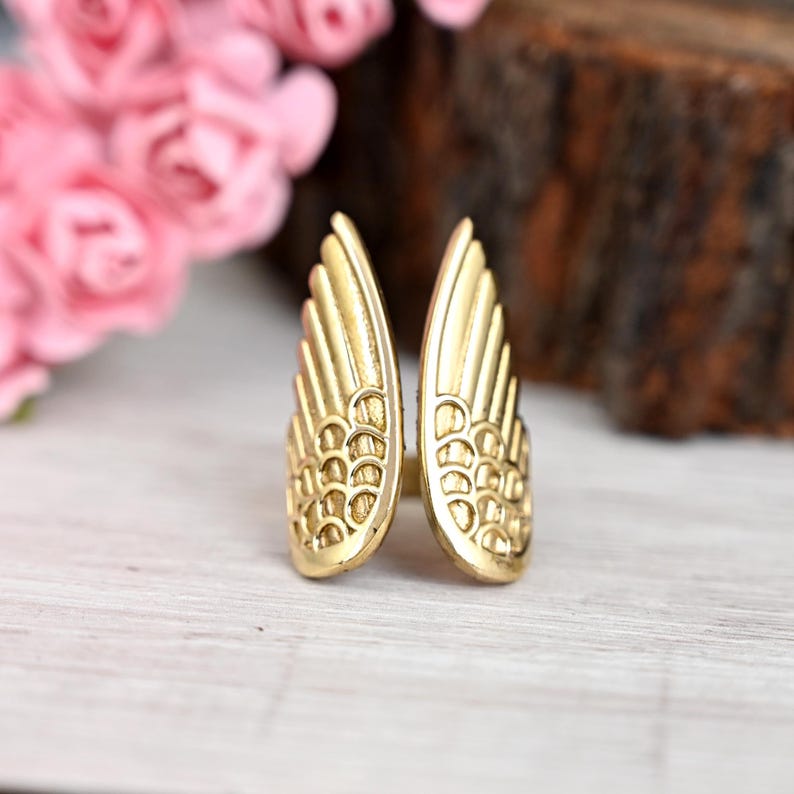 K&ouml;nnte beinhalten: Ein goldfarbener Ring in Form von ausgebreiteten Fl&uuml;geln. Der Ring hat detaillierte Federmuster. Der Hintergrund ist verschwommen, mit rosa Blumen und einem Holzelement.