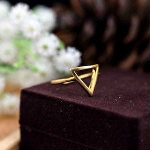 Puede incluir: Un anillo dorado con un diseño de triángulo geométrico. El anillo está sobre una superficie de terciopelo marrón oscuro.