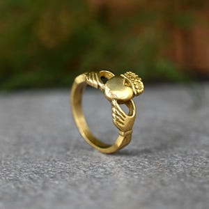May include: A gold-colored Claddagh ring with a heart design and two hands holding the heart. The ring is a traditional Irish symbol of love, loyalty, and friendship.