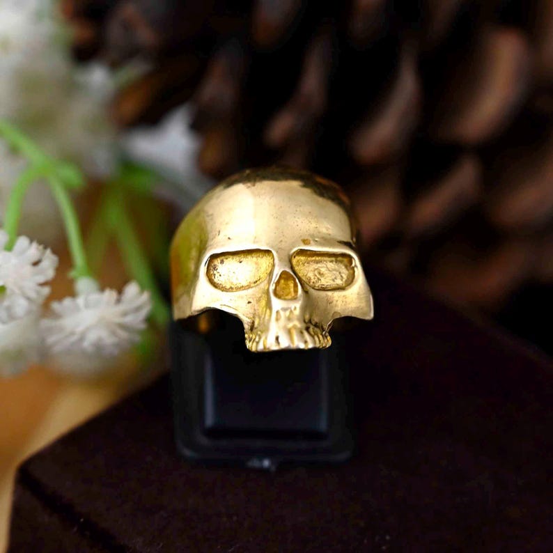 May include: A gold skull ring with a detailed design. The ring is on a black display stand.