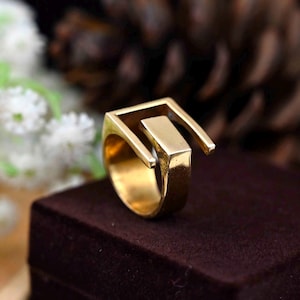 Pode incluir: Um anel dourado com um design geométrico. O anel é feito de uma única peça de metal que é dobrada em forma de quadrado com um quadrado menor cortado no centro. O anel está sobre uma superfície marrom.