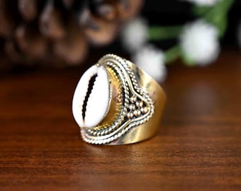 Peut inclure: Une bague en or avec un coquillage de cauris blanc au centre. La bague a une bande décorative avec un motif de perles.